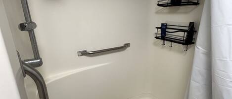 A shower with a grab bar, a handheld showerhead, and two black soap dispensers on wall-mounted shelves.