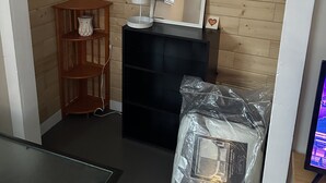 Una habitación con paredes de madera, un mueble negro, un estante de madera pequeño, un espejo y un objeto envuelto en plástico en el suelo.