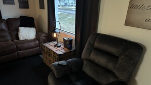 Una sala de estar con un sofá de cuero marrón, un sillón reclinable, una maleta antigua y una mesita con una lámpara y un altavoz.