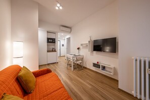A modern living room with a sofa, a dining table with chairs, a flat-screen TV mounted on the wall, and a kitchen area in the background.