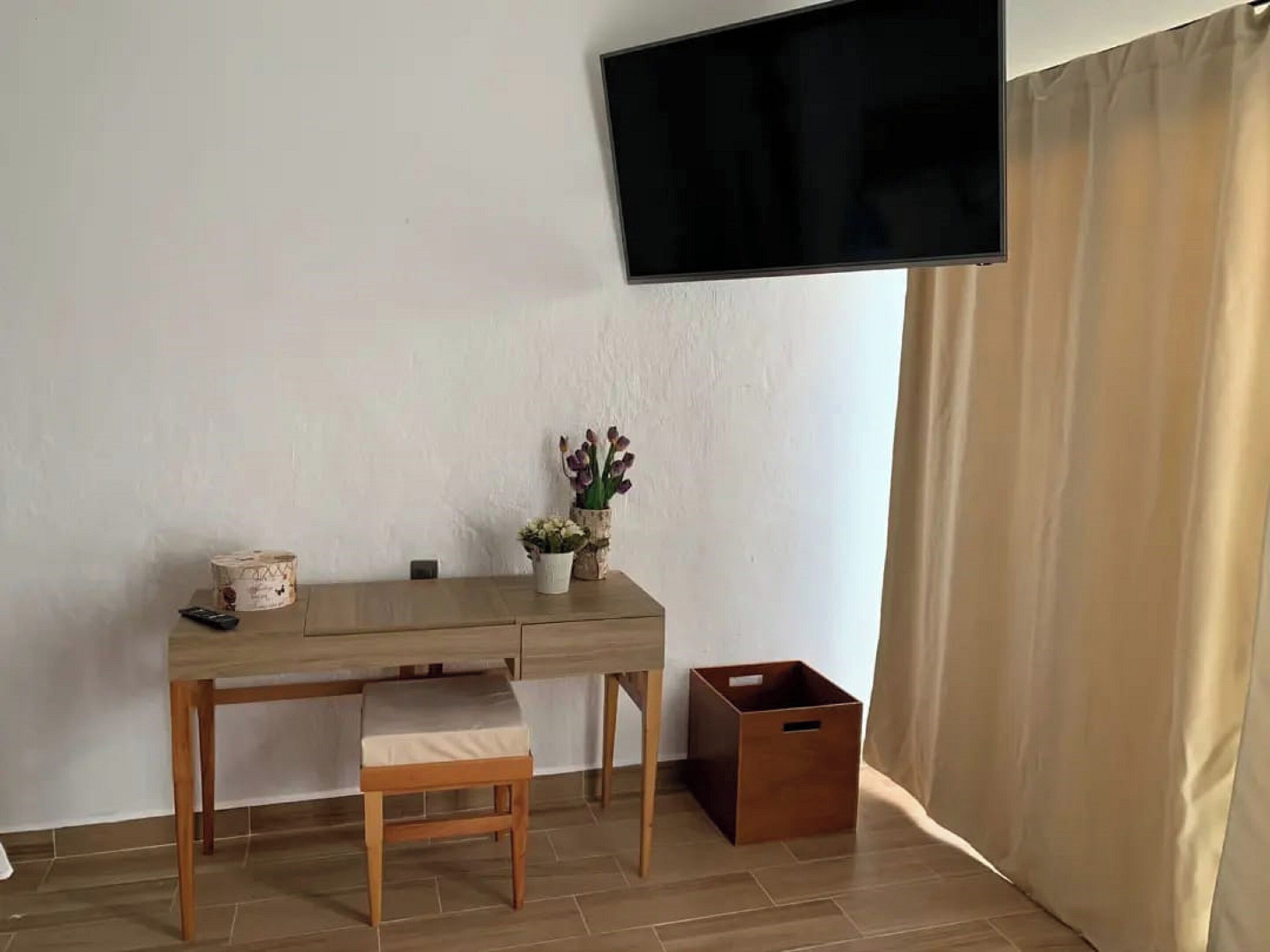 Una habitación con un escritorio de madera, un taburete pequeño, un televisor montado en la pared y un cesto de basura.