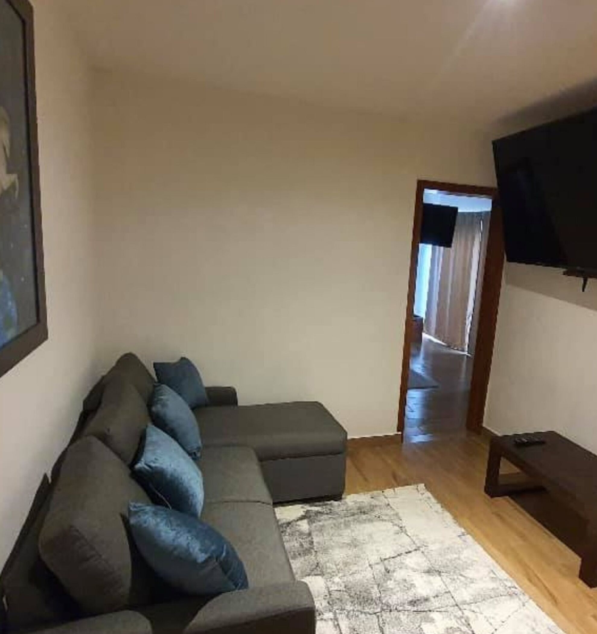 A living room with a sectional sofa, a flat-screen TV mounted on the wall, and a wooden floor.