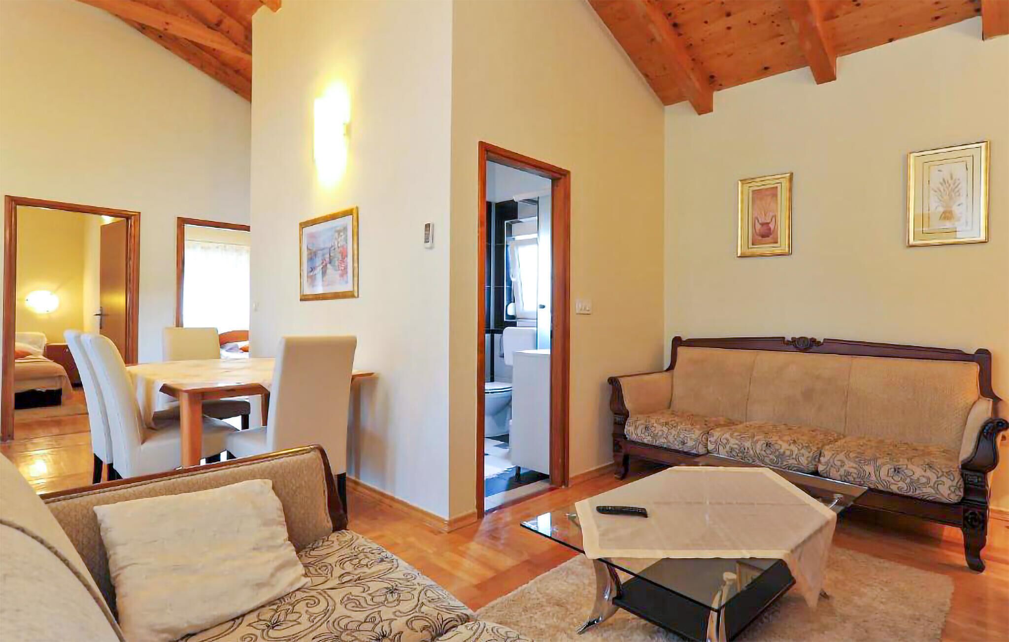 Uma sala ampla com sofá, mesa de jantar com cadeiras e uma mesa de centro de vidro. Há uma porta que dá para um banheiro.