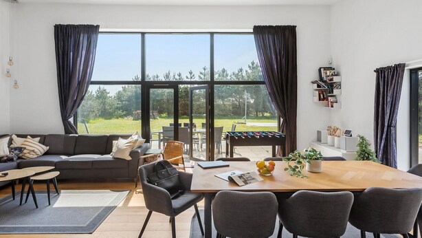 Una sala moderna con sofá, mesa de comedor, sillas y mesa de futbolín. Amplias ventanas ofrecen vistas al exterior.