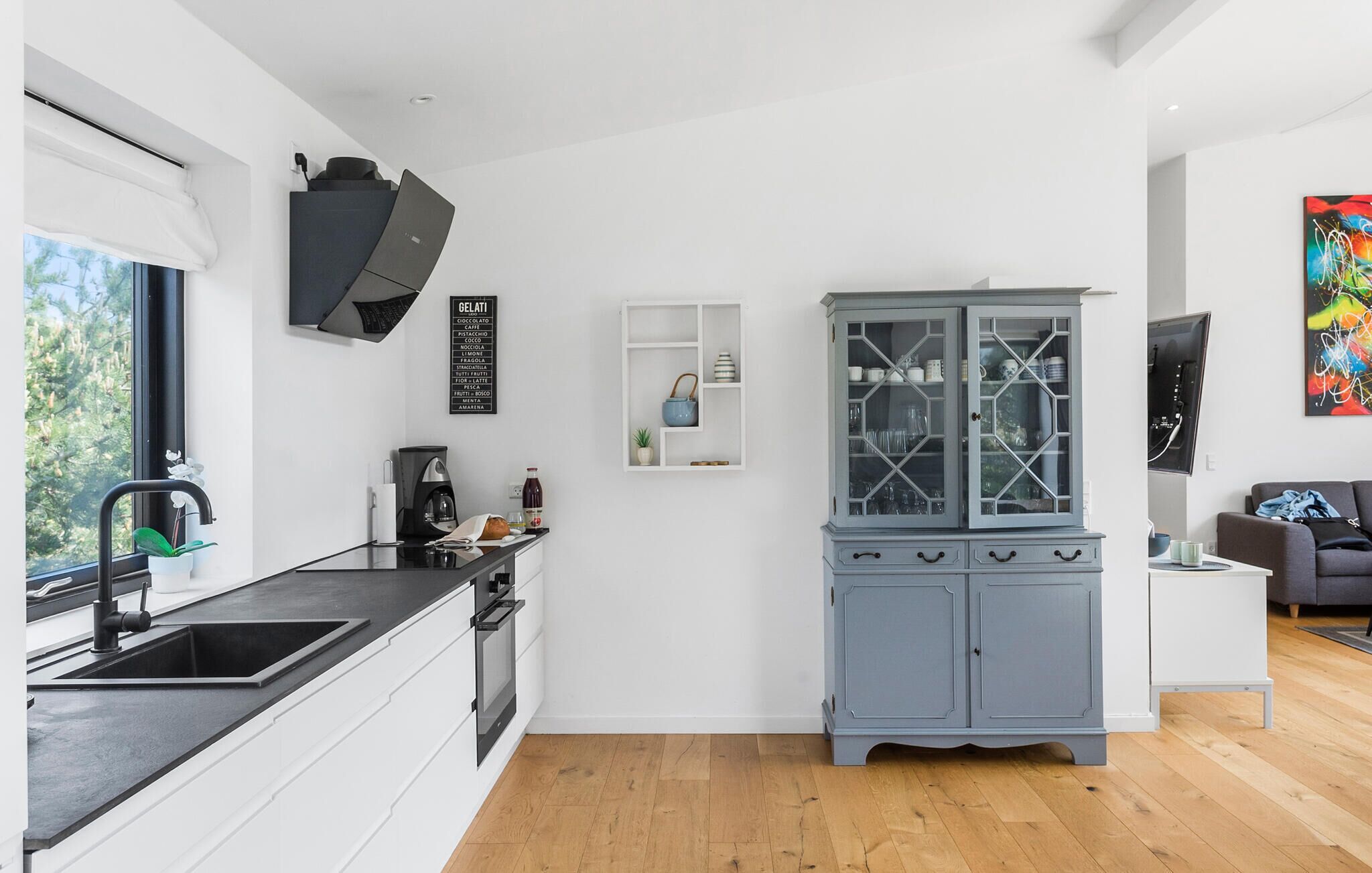 Una cocina moderna con horno empotrado, cafetera y lavamanos. Se observa un mueble con puertas de vidrio y una pequeña mesa blanca con una lámpara.