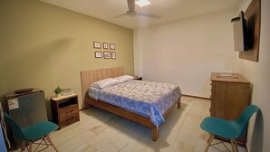 Un dormitorio con cama, escritorio, dos sillas, ventilador de techo y cuadros en la pared.
