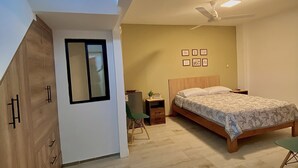 Un dormitorio con cama, una silla, una mesita, ventilador de techo y ventana.