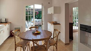 A dining area with a round wooden table, four wooden chairs, a window with a view of a garden, and a kitchen area with white cabinets and a built-in oven. - Les Lauriers De Fontvieille (Fonvieille)