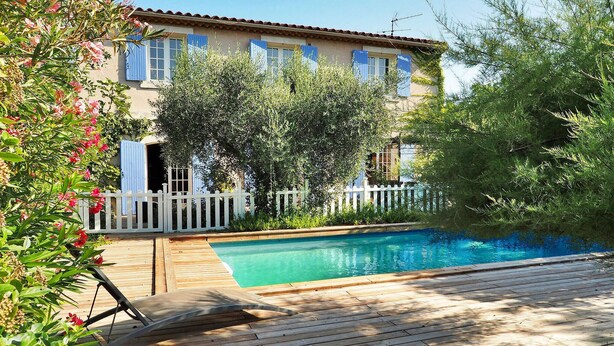 A poolside area with a wooden deck, a pool, and a building with blue shutters and a white fence. - Les Lauriers De Fontvieille (Fonvieille)