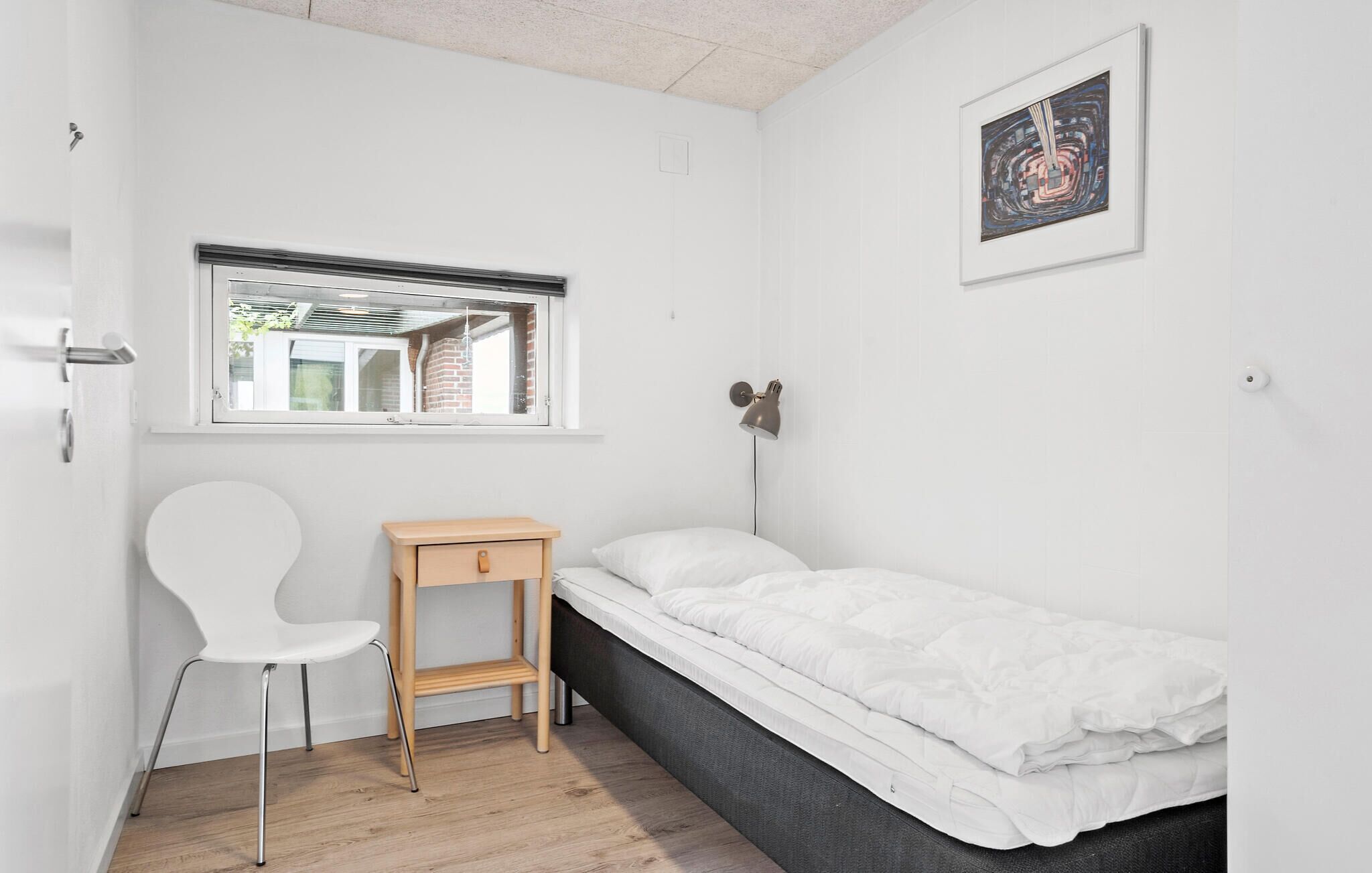 Une petite pièce blanche, avec un lit simple, une table de chevet en bois, une chaise blanche, une fenêtre et une lampe fixée au mur.