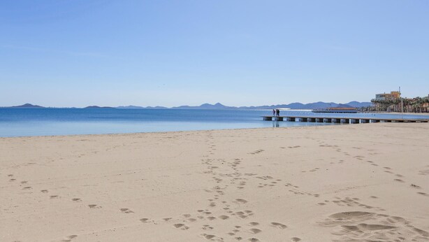 Una spiaggia con impronte, un molo e montagne in lontananza.
