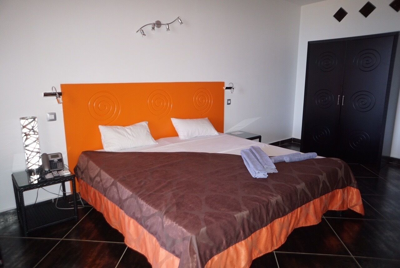 A hotel room with an orange headboard, a bed with a brown and orange bedspread, a nightstand with a telephone, and a black wardrobe.