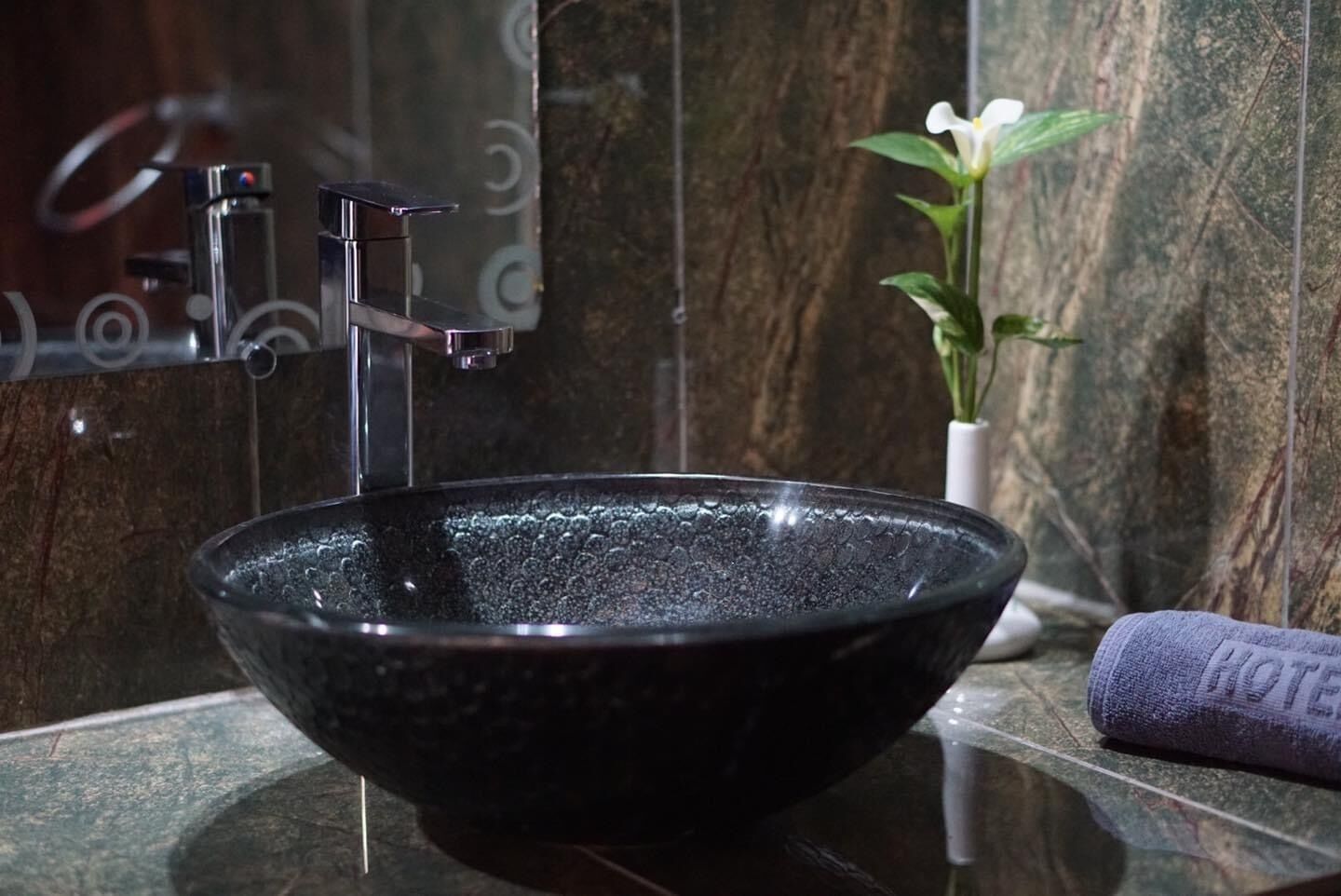 A black vessel sink with a modern faucet in a bathroom with marble walls and a potted plant.