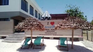 A beachside hotel with a signboard, two thatched umbrellas, and three green lounge chairs. - Hotel de la Poste (Antsiranana)