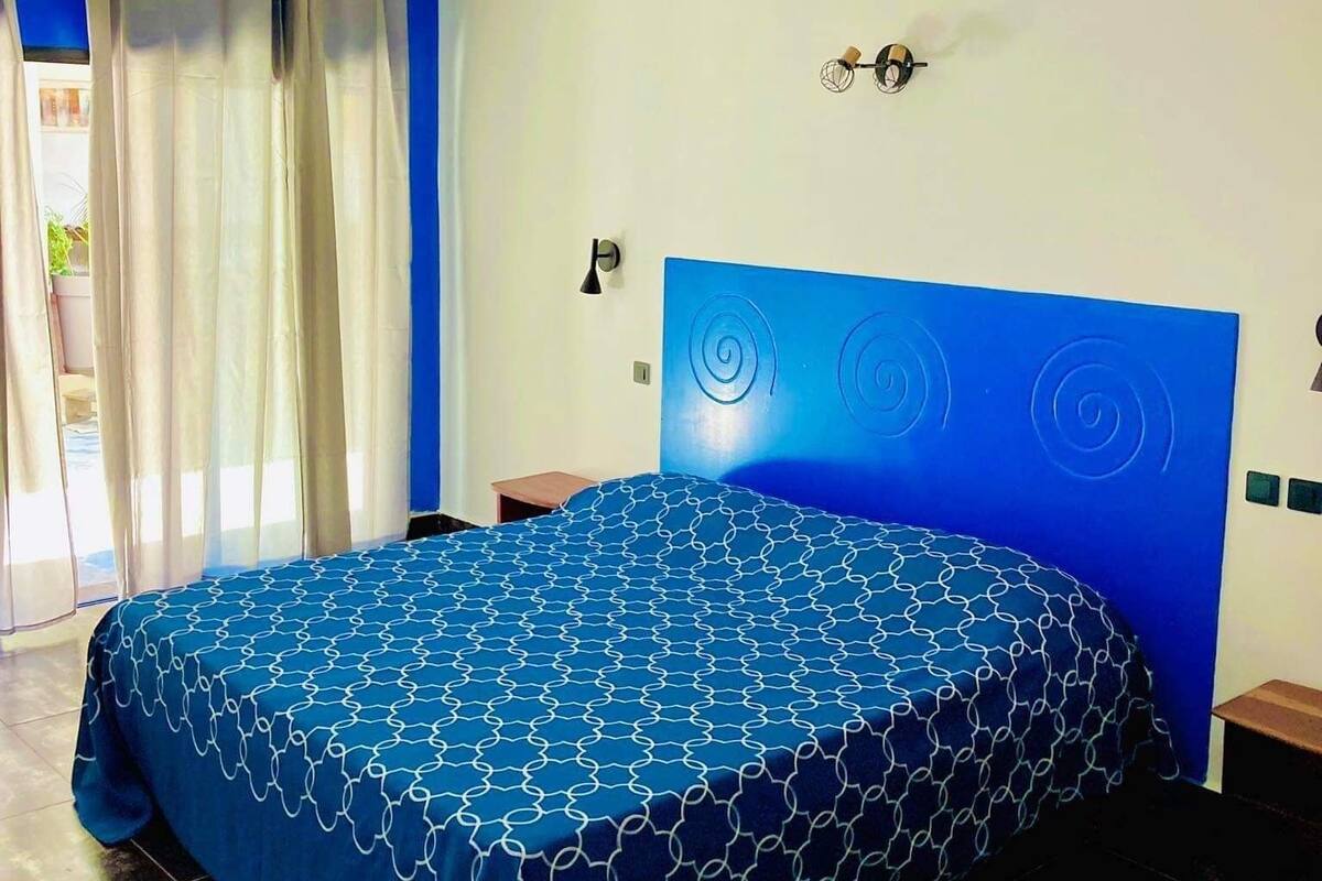 Een slaapkamer met een blauw bed met een patroon, een hoofdbord met een rond ontwerp, een nachtkastje en een raam met gordijnen.