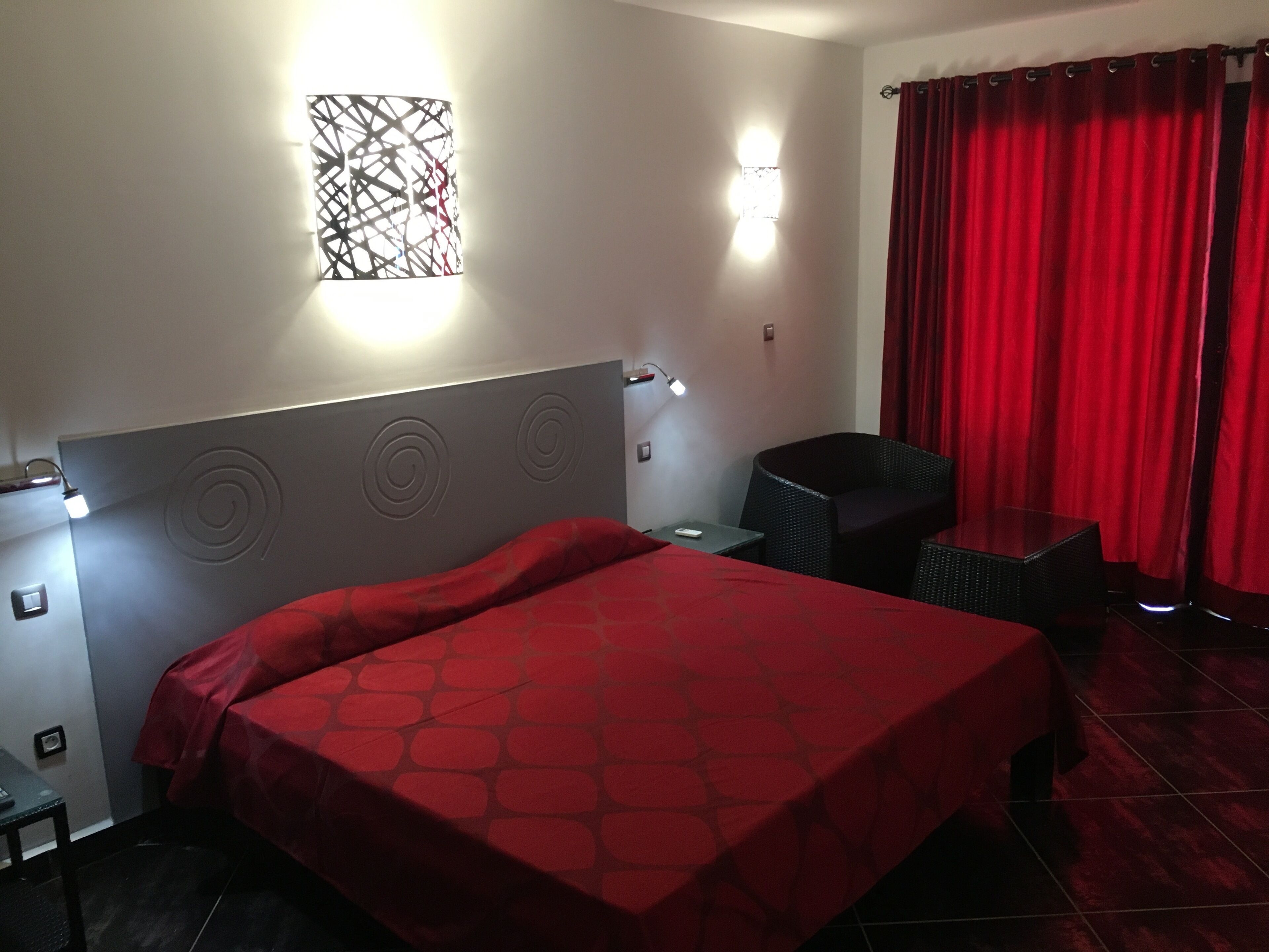 A hotel room with a bed, a chair, a small table, a wall-mounted light, and a red curtain.