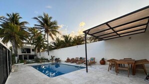 A swimming pool with lounge chairs and a wooden table, surrounded by palm trees and a white wall. - Mizu Villa (Chelem)