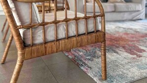 Una cadira de vimet amb respatller i seient amb amortidor, col·locada sobre un focarro estampat en un saló amb un sofà i un quadre a la paret.