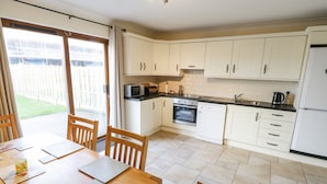 Een keuken met witte kasten, een magnetron, oven en vaatwasser. Er is een eettafel met stoelen en een raam met gordijnen.
