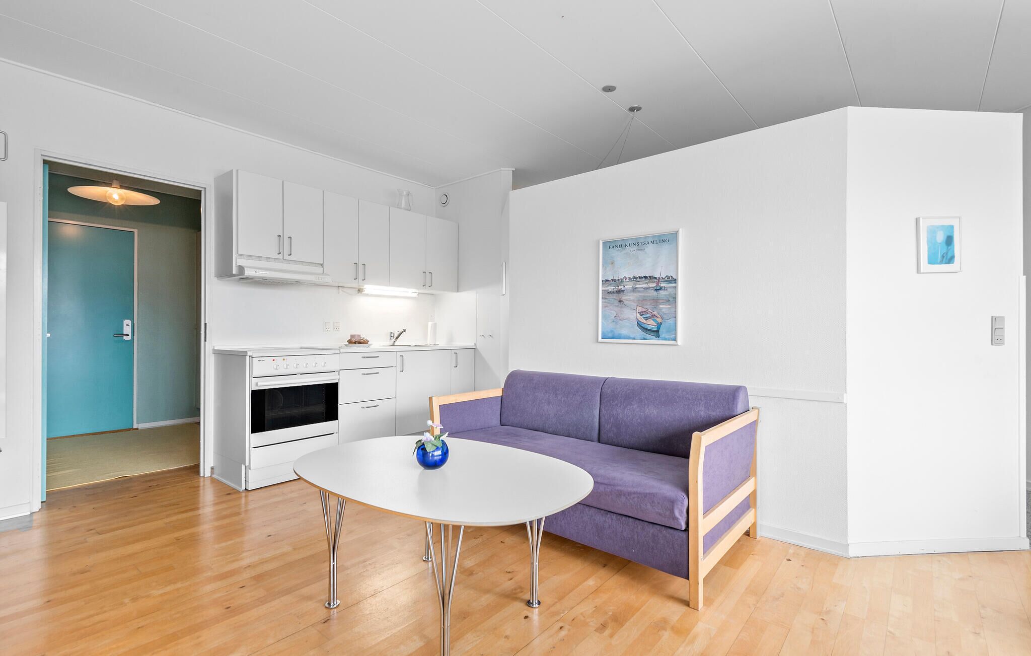Une cuisine moderne avec des meubles blancs, un four encastré et un coin repas comprenant une table ronde et un canapé violet.