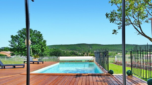 Une piscine rectangulaire dotée d’une terrasse en bois et de rampes en métal, entourée d ’une clôture et d’arbres, avec un paysage vallonné en arrière-plan.