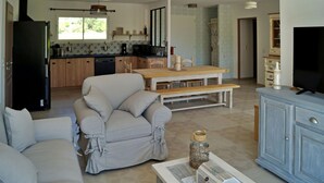 Un salon avec un canapé, une table basse et un téléviseur. Au fond, on aperçoit une cuisine avec une fenêtre et un évier.