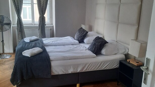 A bed with white linens, a dark blanket, and a bedside table with a lamp. - Metropolitan Charm: Stylish 2+1 Apt@ Vienna's Core (Vienna)