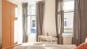 Ein Schlafzimmer mit einem Bett, einem Schreibtisch, einem Stuhl, einem Holzschrank und einem Fenster mit Vorhängen.
