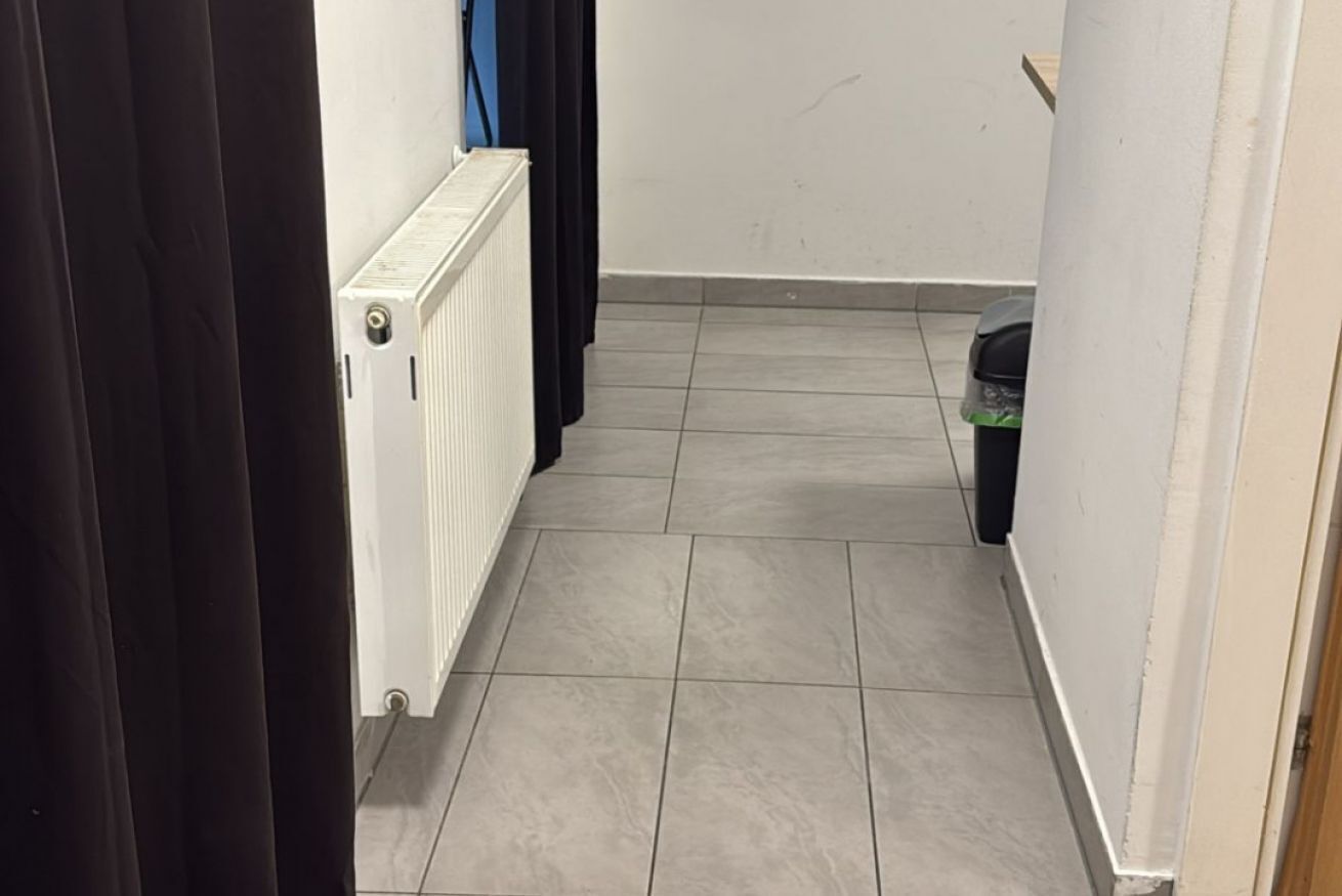 Ein schmaler Flur mit Heizkörper, schwarzem Mülleimer und Tür mit schwarzen Vorhängen.