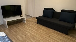 Habitación con una televisión de pantalla plana sobre un soporte blanco, un sofá negro con dos cojines y suelo de madera.