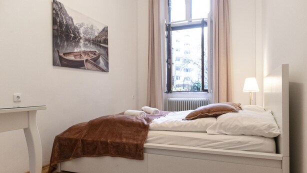 Un dormitorio con cama, ventana con cortinas, radiador, una mesa y un cuadro en la pared.