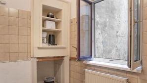 A small bathroom with a window, a radiator, and a shelf with kitchen items.