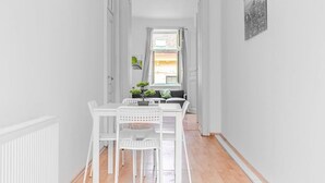 Un pasillo con una mesa y sillas blancas, una ventana con cortinas y un cuadro en la pared.