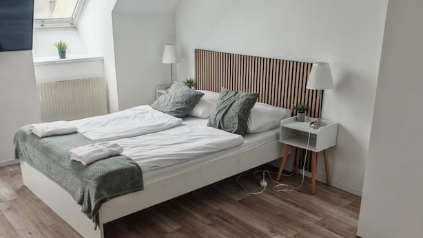 A modern bedroom with a white bed, a bedside table with a lamp, a radiator, and a small plant on the window sill.