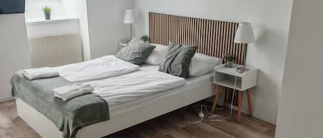 A modern bedroom with a white bed, a bedside table with a lamp, a radiator, and a small plant on the window sill.