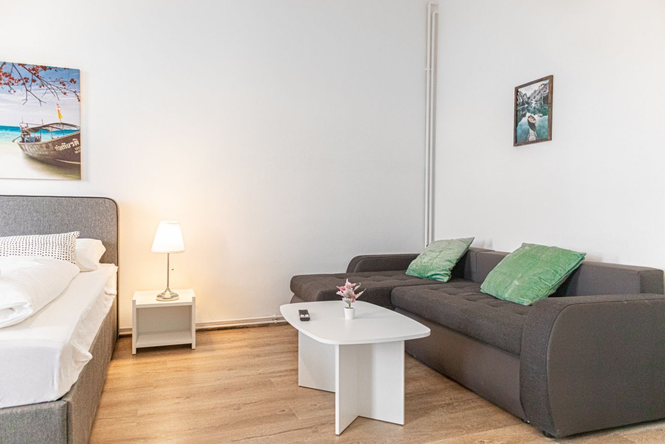 A modern hotel room with a bed, a grey sofa, a small white table, a nightstand, and a lamp.