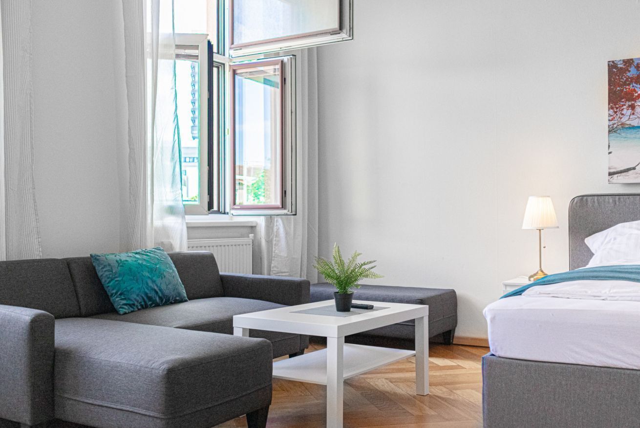 Une chambre moderne avec un canapé gris, une table basse blanche, un lit et une fenêtre avec des rideaux.