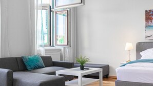 A modern room with a grey sofa, a white coffee table, a bed, and a window with curtains.
