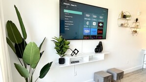 A modern living room with a mounted TV displaying a home screen, a potted plant, a pair of glasses, and a small shelf with decorative items. - Cozy King 1BR w/ Pool + Gym Near EWR & MetLife (Elizabeth)
