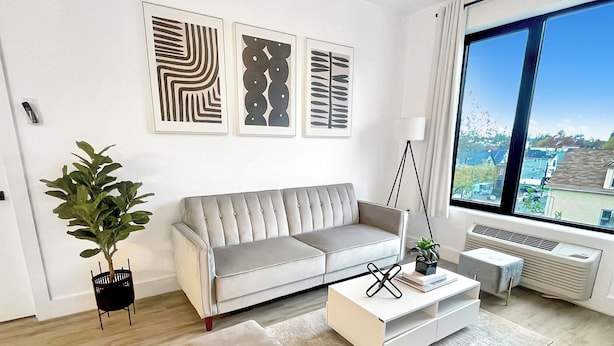 A modern living room with a grey sofa, a white coffee table, and a large window offering a city view. - Cozy King 1BR w/ Pool + Gym Near EWR & MetLife (Elizabeth)