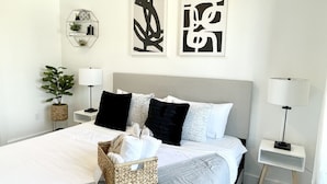 A neatly arranged bedroom with a bed, pillows, a bedside lamp, a small plant, and artwork on the wall. - Cozy King 1BR w/ Pool + Gym Near EWR & MetLife (Elizabeth)