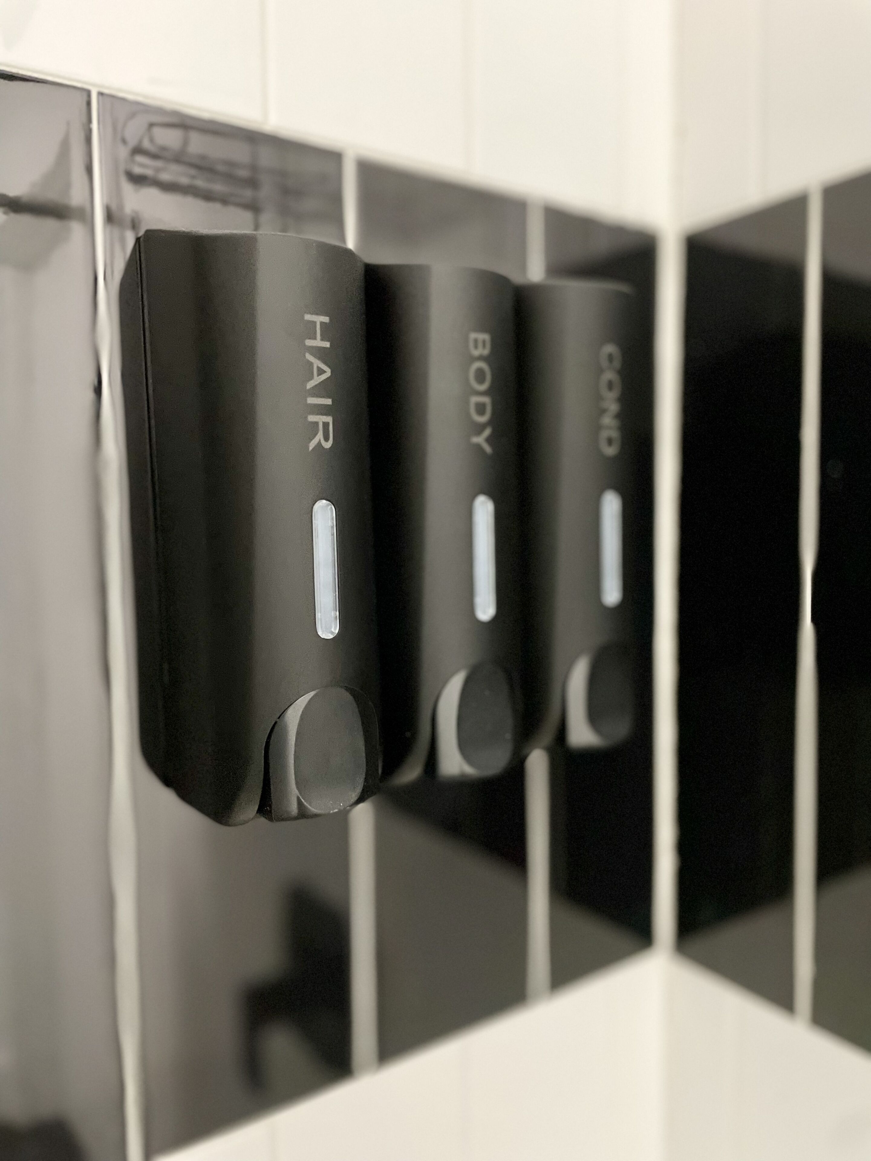 A row of black, rectangular dispensers labeled "HAIR," "BODY," and "COND" mounted on a tiled wall.