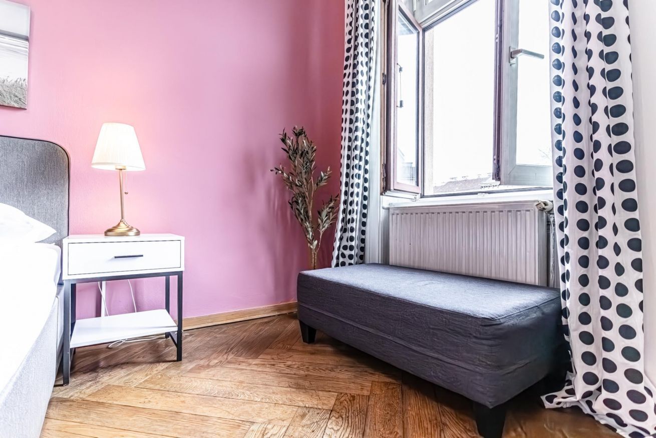 Een kamer met een bed, nachtkastje met lamp, een bankje, een raam met gordijnen en een radiator.