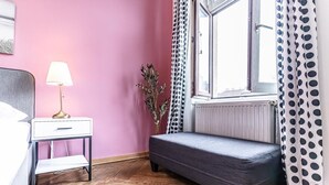 A room with a bed, bedside table with a lamp, a bench, a window with curtains, and a radiator.