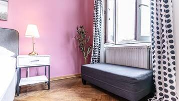 Een kamer met een bed, nachtkastje met lamp, een bankje, een raam met gordijnen en een radiator.