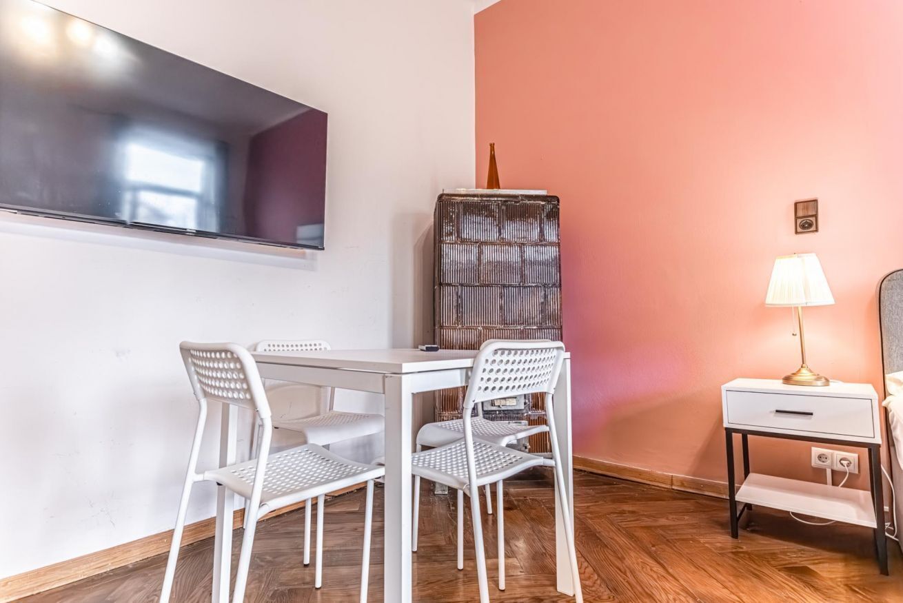 Een kamer met een witte tafel en stoelen, een aan de muur bevestigde televisie, een hoog decoratief object, een lamp en een nachtkastje.