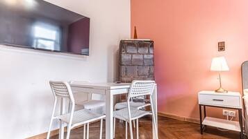 Een kamer met een witte tafel en stoelen, een aan de muur bevestigde televisie, een hoog decoratief object, een lamp en een nachtkastje.