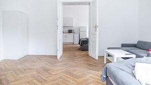 A room with a herringbone floor, a bed, a grey sofa, a white coffee table, and an open door leading to a kitchen area with cabinets and appliances. - Classy 2BR Apt. w/ Exclusive Location near AKH (Wien)
