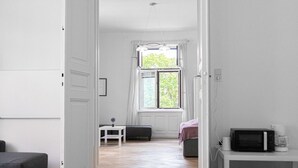 Una habitación con una cama, una mesita con una lámpara, una ventana con cortinas y un microondas en un estante.