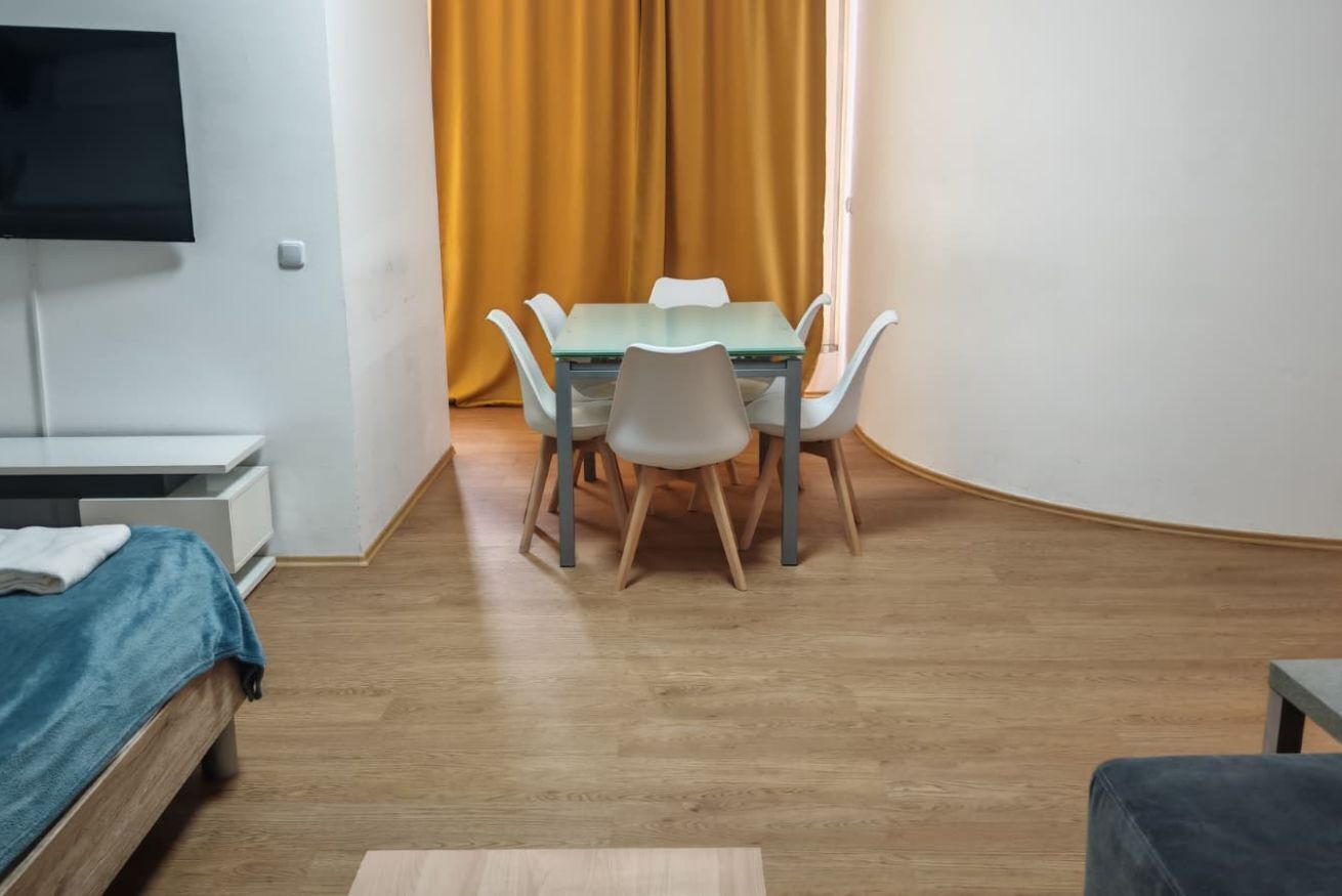 Ein Zimmer mit einem Bett, einem an der Wand montierten Fernseher, einem Esstisch mit Stühlen und einem Fenster mit Vorhängen.